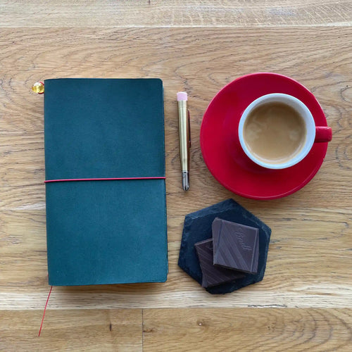 Bielinis handmade traveler’s journal Atlas in petrol blue leather, flat lay with espresso and dark chocolate on wooden table.