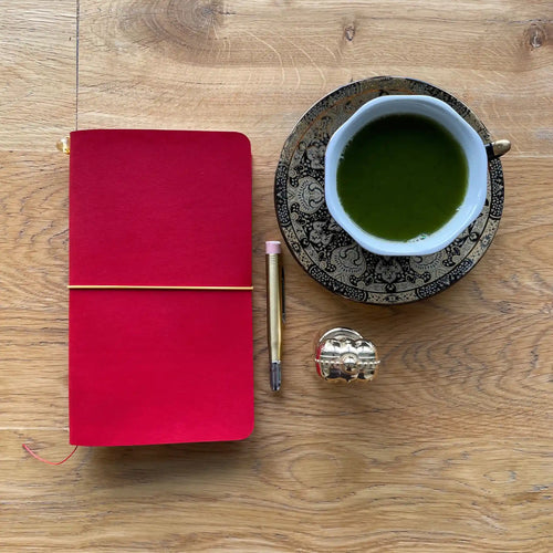 Bielinis handmade traveler’s journal Atlas in red leather, flat lay with matcha tea and Christian Clive perfume on wooden table.